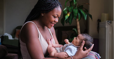 A woman smiles at a baby in her arms, surrounded by a cozy indoor setting with plants and soft lighting.