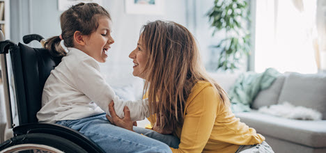 A woman and a girl in a wheelchair share a joyful moment, smiling and interacting in a cozy living room.