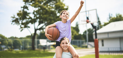 A young child is playing basketball, holding a ball above a hoop against a blue sky with clouds.