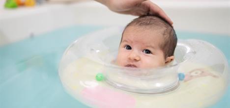 A baby in a floating bath ring looks up, with a hand gently touching their head, surrounded by water.