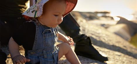 A young child in a colorful hat sits on a rocky surface, focused on something in front of them, with a soft sunset glow in the background.