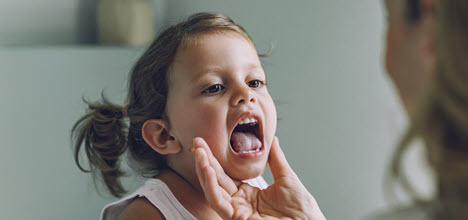 A young girl is having her mouth examined by an adult, with a focus on her open mouth and playful expression.
