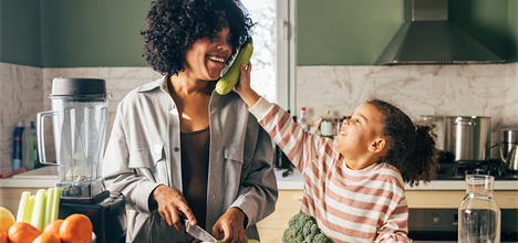 A woman and a child playfully interact in a kitchen, with the child holding a cucumber while the woman smiles and prepares food.