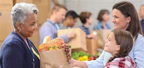 A woman hands a bag of groceries to another woman and a girl, smiling in a community food distribution setting.