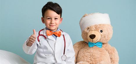 A child in a doctor's coat gives a thumbs up, sitting next to a teddy bear with a bandaged head.