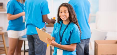 A group of volunteers in blue shirts, with one girl smiling and holding a clipboard, while others work in the background.