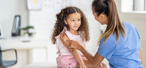 A young girl with curly hair sits on a medical table, smiling at a nurse in scrubs who is applying a bandage to her arm.
