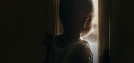 A child gazes out a window, illuminated by soft light, with a contemplative expression. The scene is dimly lit, creating a moody atmosphere.