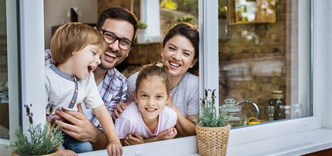 A happy family of four smiles and leans out of a window, with potted plants on the sill.