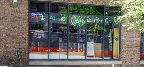 A storefront with large windows displaying neon signs for a dispensary, featuring products like Delta 8, Kratom, and CBD.