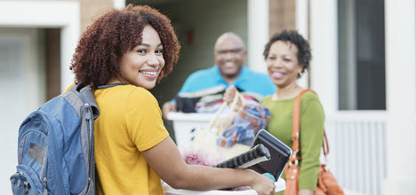 A young woman smiles while carrying a basket of items, accompanied by two adults, outside a building.