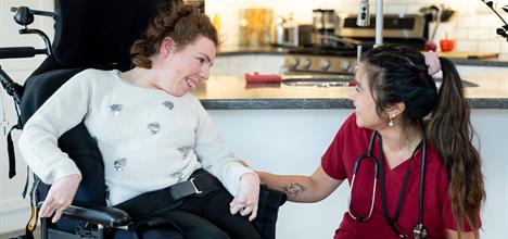 A caregiver interacts warmly with a young woman in a wheelchair, both smiling in a bright kitchen setting.