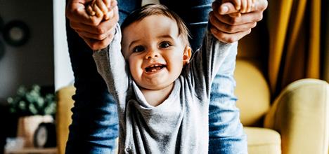 A joyful baby is being held by the hands, smiling widely. The background features a cozy living space with soft furnishings.
