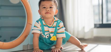 A toddler in colorful clothing crawls on a wooden floor, with sunlight streaming through a window in the background.
