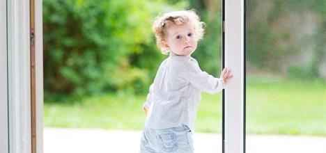 A young child with curly hair stands by an open door, looking back with a curious expression. Lush greenery is visible outside.
