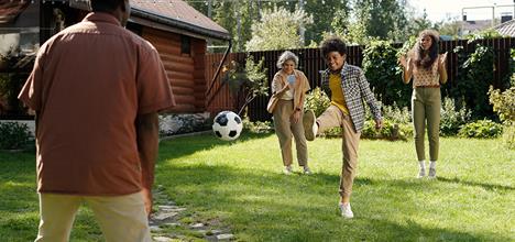A group of four people play soccer in a sunny backyard, with one person kicking the ball while others watch and cheer.