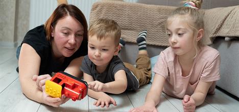 A woman and two children are playing on the floor with a toy vehicle, focused and engaged in their activity.