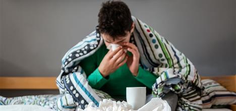 A person wrapped in a striped blanket sits on a bed, holding a tissue and surrounded by used tissues and a cup.