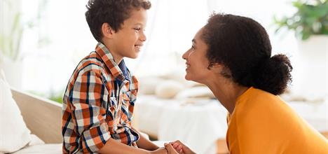 A woman and a boy share a joyful moment, smiling at each other while holding hands in a bright, cozy room.