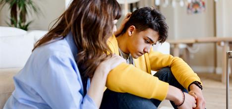 A young man sits on the floor, looking down, while a woman comforts him with a hand on his shoulder. They are in a cozy, modern room.