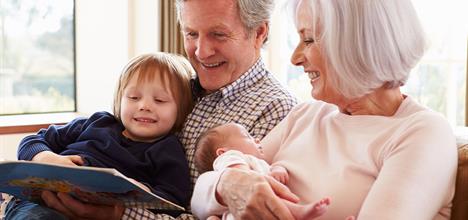 A family scene with a child reading a book to grandparents, who are smiling and holding a baby. Bright, cozy indoor setting.