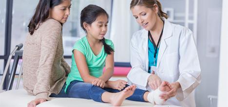 A doctor is examining a young girl's injured foot while her mother watches attentively in a medical setting.