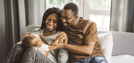 A happy couple sits on a couch, playing with their baby. They share smiles and affection in a cozy, well-lit room.