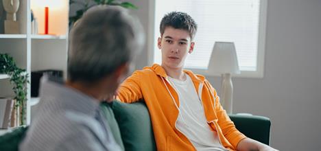 A young man in an orange hoodie sits on a couch, engaged in conversation with an older man, in a cozy, well-lit room.