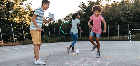 Three children are playing on a basketball court, jumping and laughing. They are using chalk to draw on the ground.